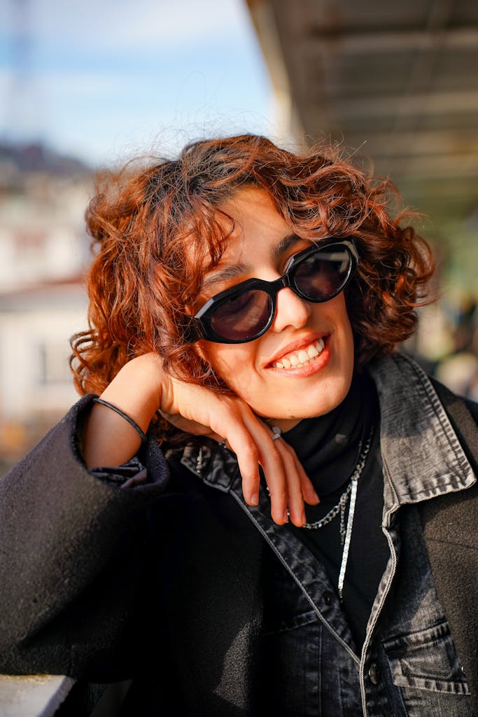 A stylish woman with curly hair and sunglasses smiling outdoors.