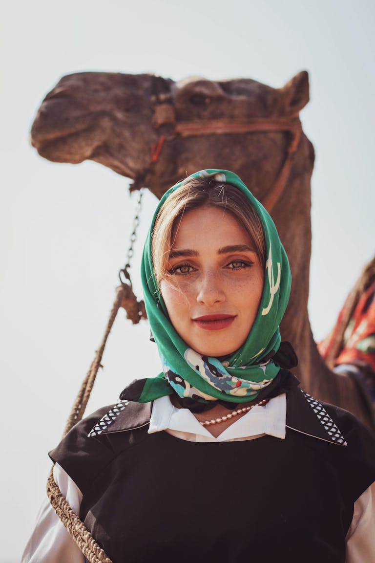 A woman in a headscarf stands confidently beside a camel under the bright desert sun.