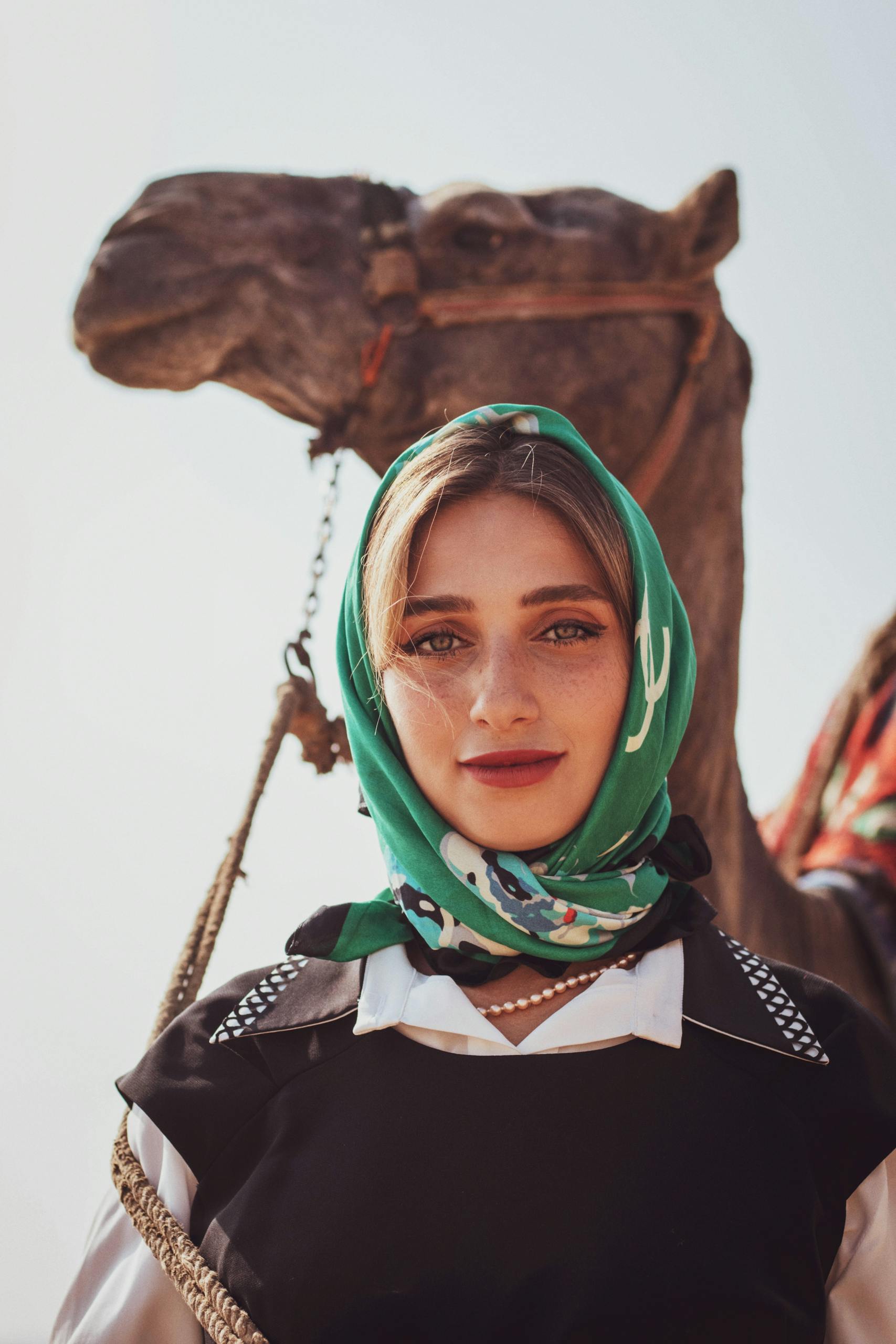 A woman in a headscarf stands confidently beside a camel under the bright desert sun.