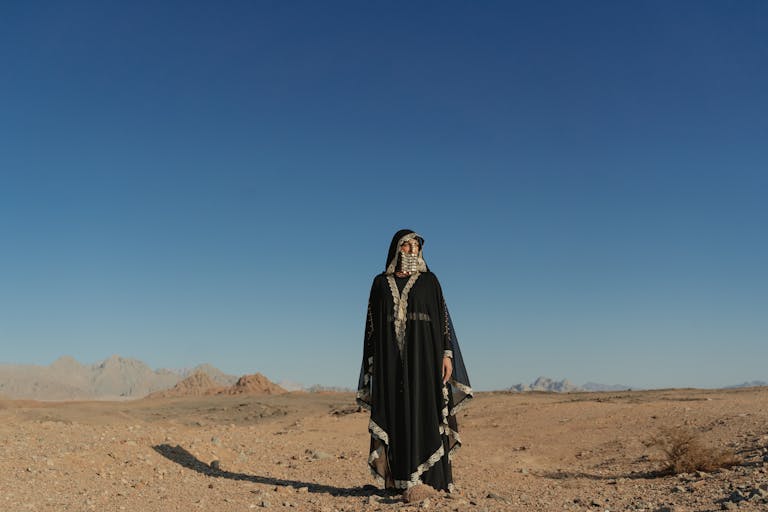 A woman stands alone in traditional attire in a vast desert with a blue sky.