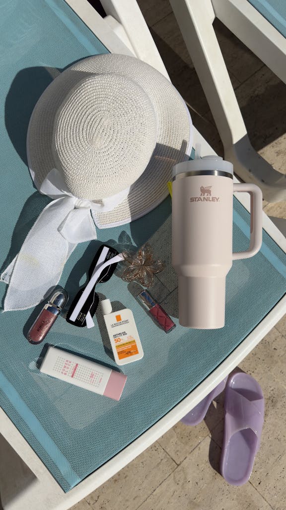 Flat lay of summer accessories on a sunbed, featuring a hat, sunscreen, and stylish bottle.